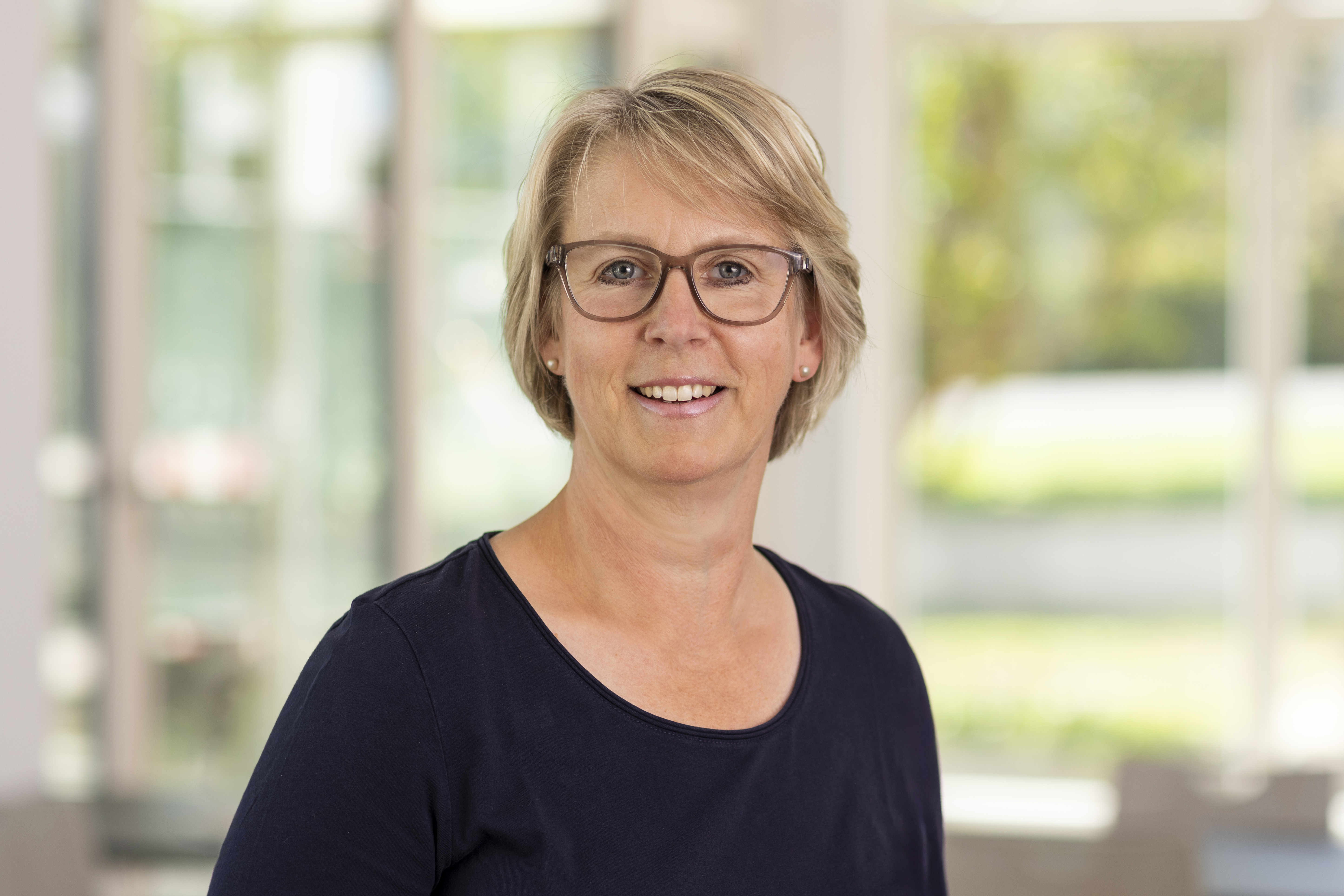 Porträt einer lächelnden Frau mit kurzen blonden Haaren und Brille in einem dunklen Oberteil vor hellem, unscharfem Hintergrund.
