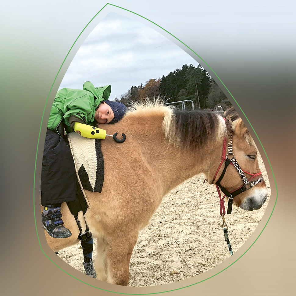 Der 9-jährige Luca trägt eine grüne Jacke und liegt entspannt auf dem Rücken eines Pferdes.