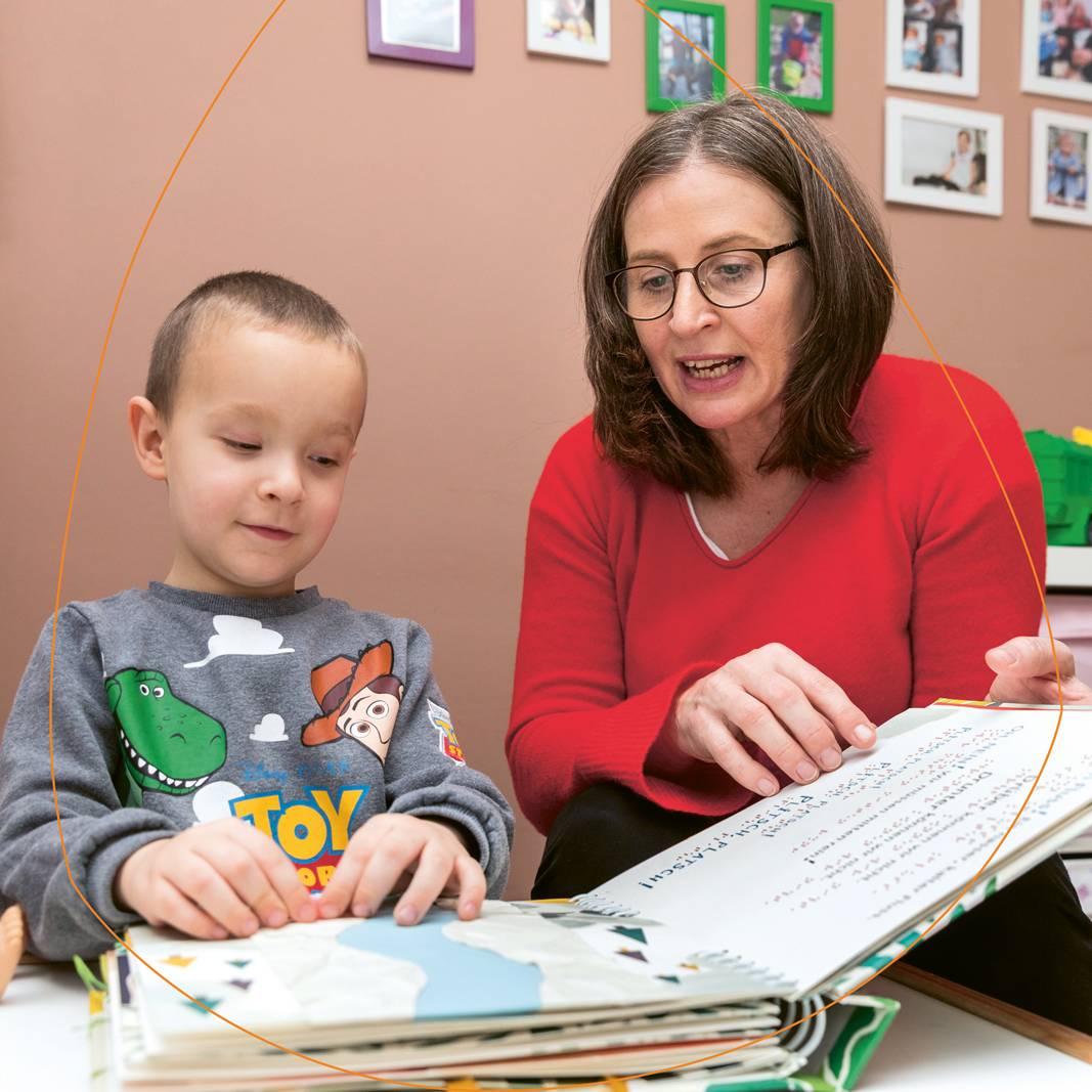 Maria Wiesner, eine Frau mit rotem Oberteil, schaut mit Maks, einem blinden Jungen im Toy-Story-Pullover, ein Buch an.