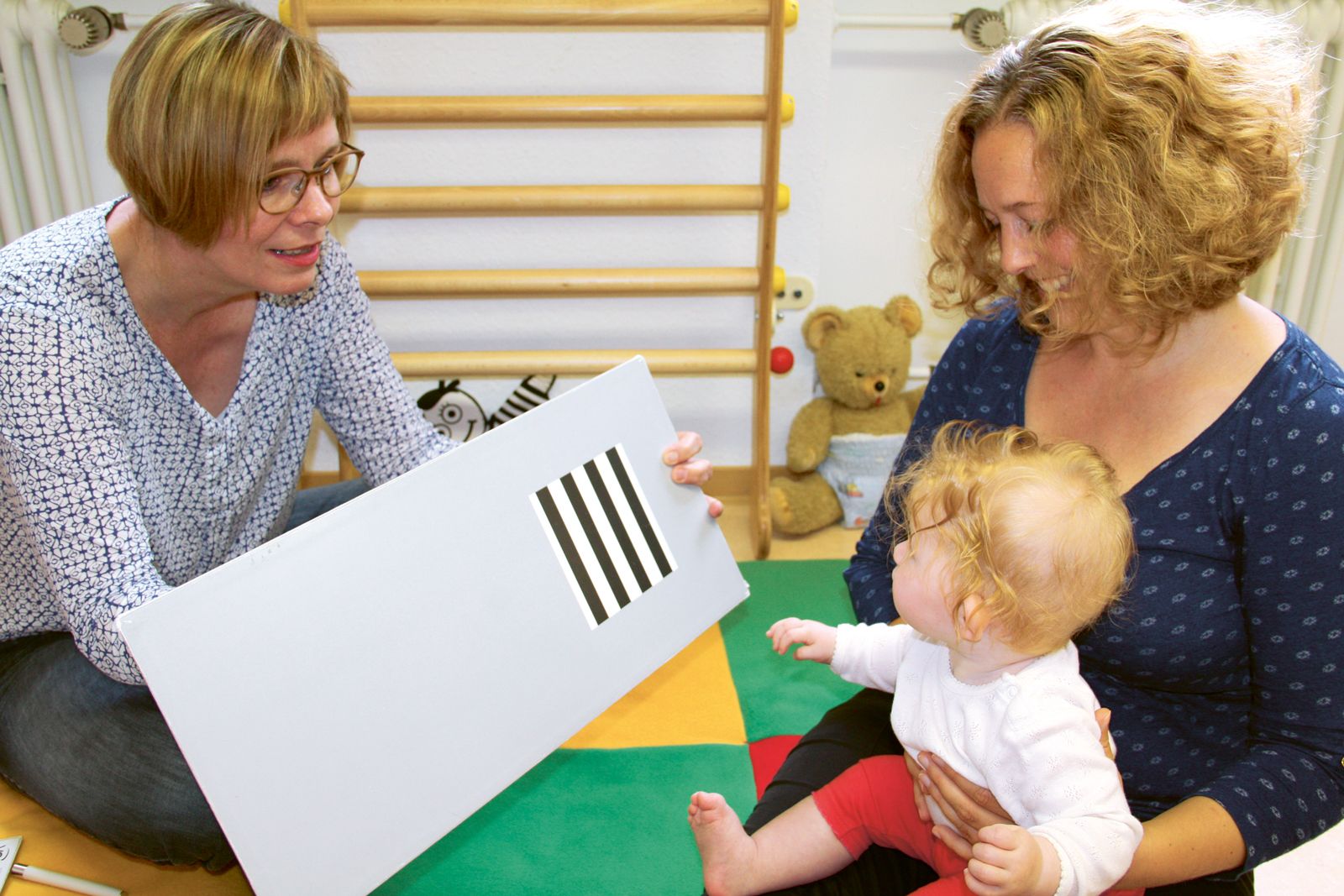 Eine Frau führt mit einem Kleinkind, welches auf dem Schoß seiner Mutter sitzt, einen Sehschärfte-Karten-Test durch.