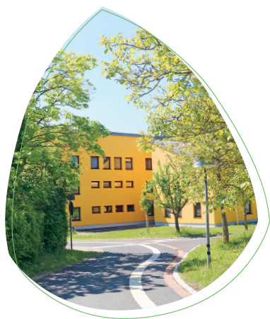 Hier ist ein Bild des Blindeninstituts Würzburg an einem sonnigen Tag. Das Gebäude ist von vielen grünen Bäumen und Wiesen umgeben.
