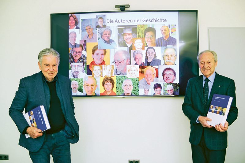 Hier ist ein Bild der beiden Herausgeber Dr. Hans Neugebauer und Dr. Wolfgang Drave mit ihrem Buch zu sehen. Im Hintergrund ist ein Bildschirm aufgestellt, auf dem die Autoren der Geschichte mit Bild dargestellt sind.