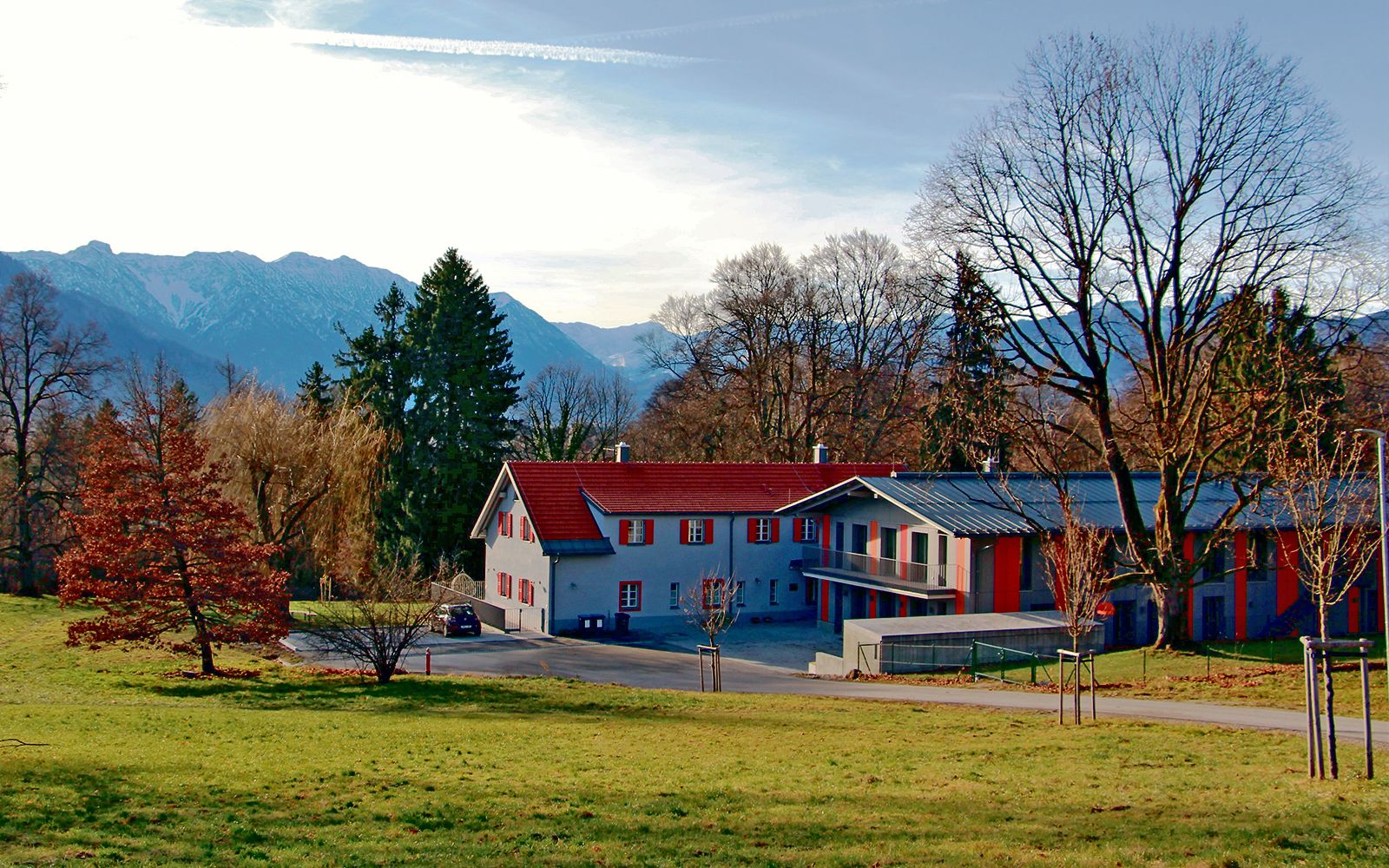 Ein Foto des Isle-Erl-Hauses, welches mitten in der Natur steht. Das Gebäude besteht aus zwei länglichen, mehrstöckigen Häusern, welche aneinandergebaut sind. Es ist von großen Bäumen umgeben und im Hintergrund erheben sich Berge.