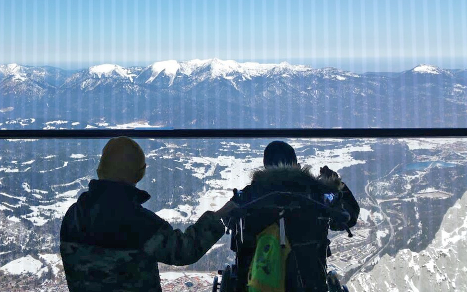 Zwei Personen, wurden von hinten fotografiert, als sie an einer Scheibe eines Ausblickpunktes auf einem Berg stehen. Vor den Personen erstreckt sich eine weite Landschaft, an dessen hinterem Ende hohe Berge aufragen.