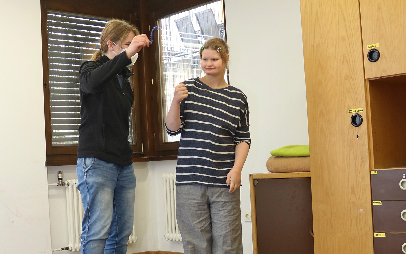 Adea steht mit Gruppenmitarbeiterin Katrin Warzlberger in einem Raum. Katrin zeigt Adea etwas in ihrer Hand und die Situation ist ruhig und friedlich.
