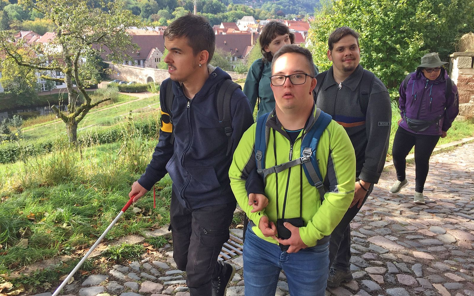 Eine Gruppe junger Menschen beim Wandern. Die rechte Person im Vordergrund trägt eine Brille und hat eine grüne Jacke an. Er läuft gemeinsam mit einem jungen Mann in blauer Jacke.