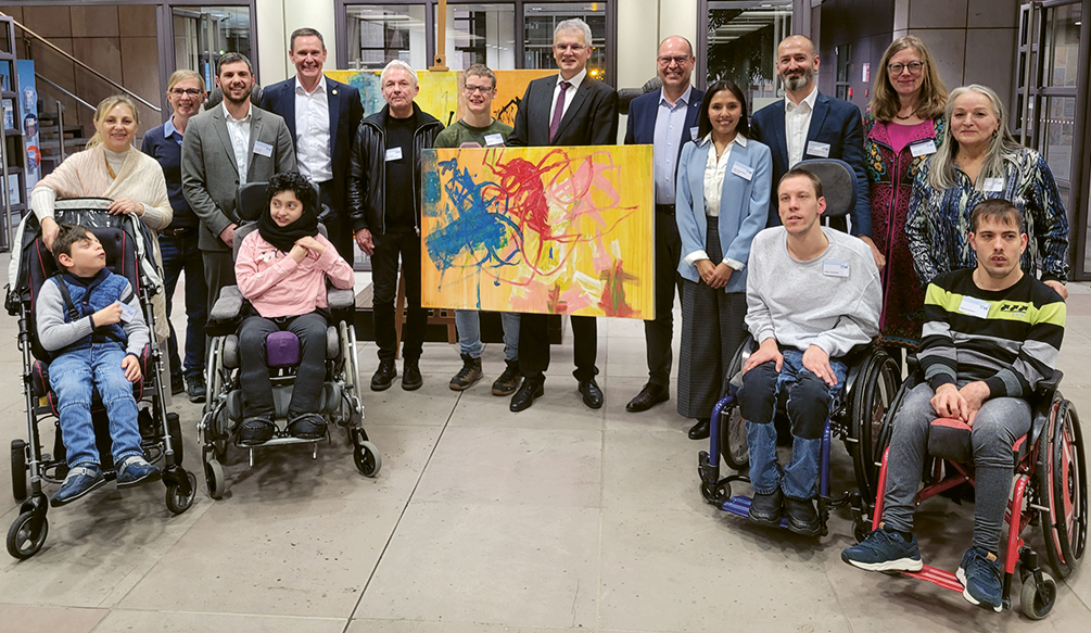 Gruppenbild, das auf der Vernissage aufgenommen wurde. Viele Menschen stehen/sitzen um ein Kunstwerk herum, das präsentiert wird. Dieses hat einen geben Hintergrund und blaue, rote, weiße und rosa Farbakzente.
