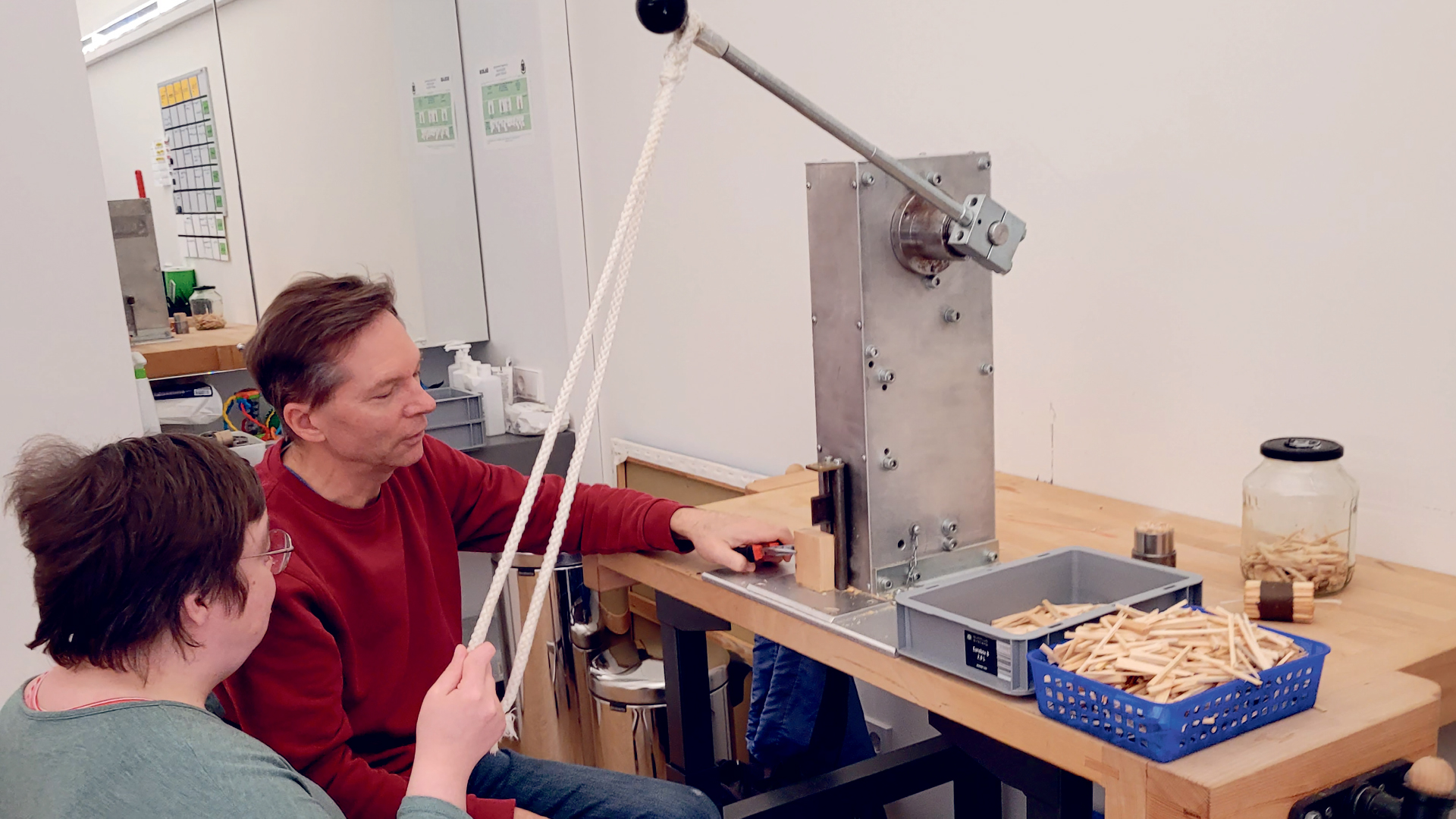 Eine Klientin sitzt vor einer Maschine, in der Holz eingespannt ist. Die Maschine wird durch einen Hebel bedient, an dem ein Seil befestigt ist. Neben ihr sitzt ein Mitarbeiter der Einrichtung. Es werden Ofenanzünder hergestellt.