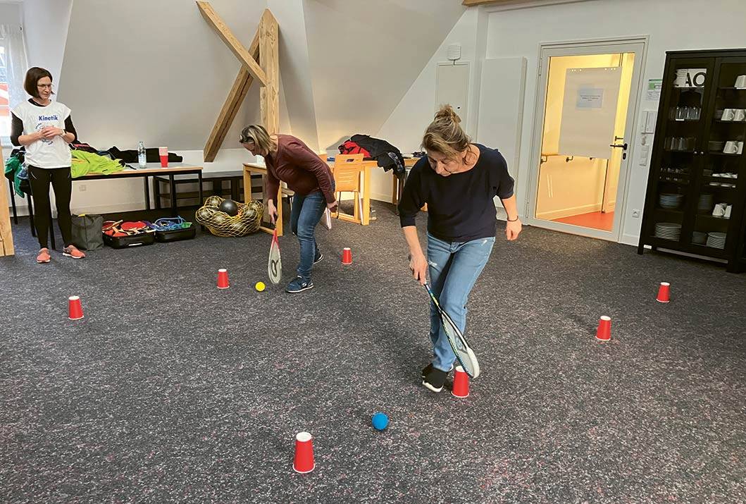 Zwei Personen, die jeweils gegeneinander Slalom um aufgestellte Bescher laufen. In der Hand haben sie jeweils einen Tennisschläger, mit dem ein blauer und ein gelber Ball durch den Parcours geführt wird.