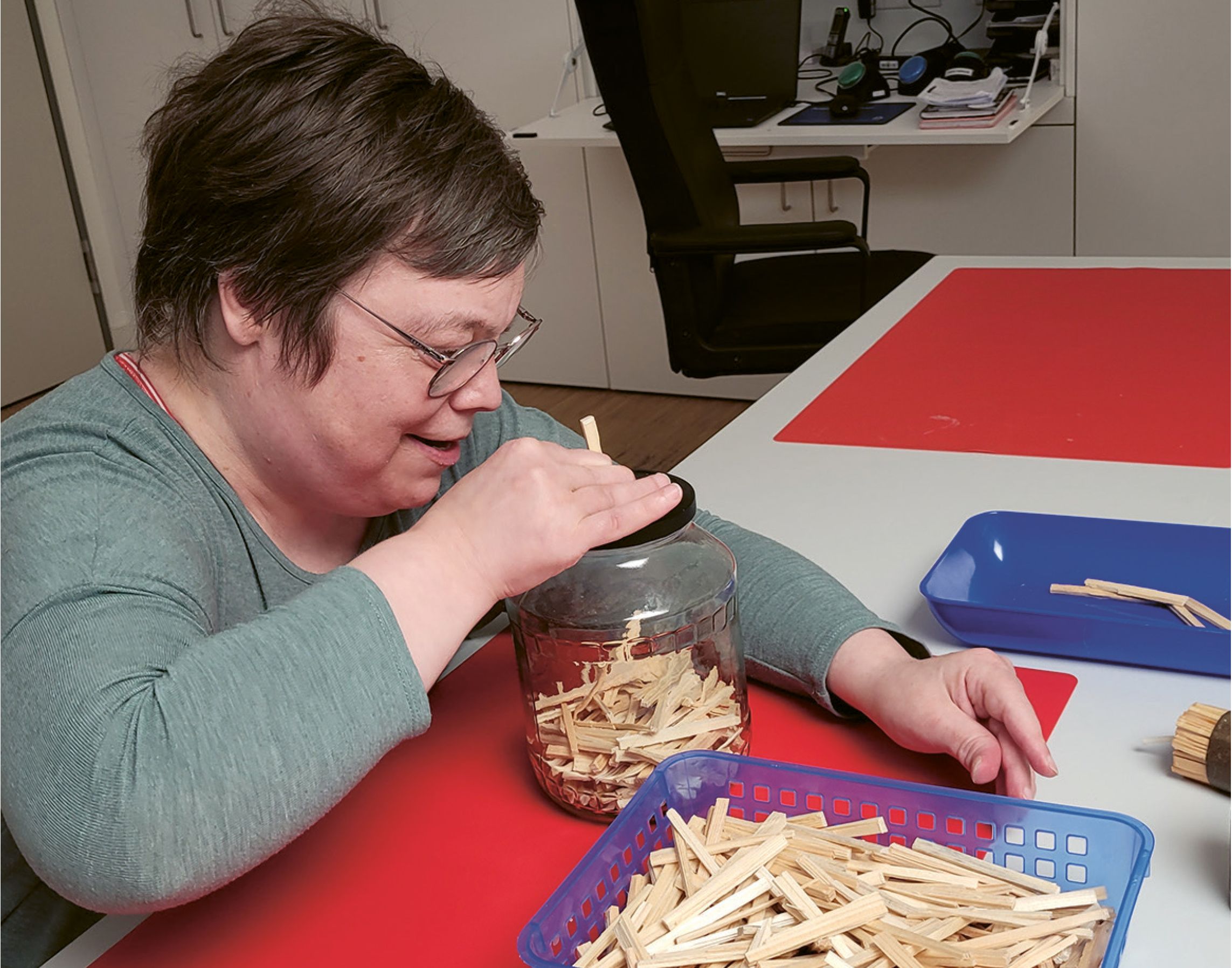 Eine Klientin prüft die Größe der Holzstücke für die Ofenanzünder, indem diese in ein Glas steckt, in dessen Deckel ein Loch ist, das die Größe vorgibt. Nicht passende Holzspalten werden aussortiert.