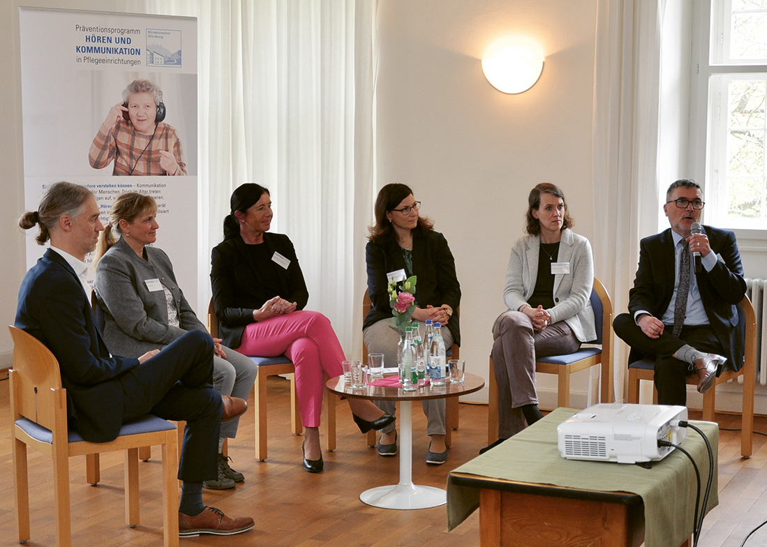 Die Teilnehmer der Podiumsdiskussion sitzen in einem Halbkreis zusammen. Johannes Spielmann (Vorstand der Blindeninstitutsstiftung) hält ein Mikrofon in der Hand und spricht. Alle anderen Teilnehmer schauen ihn an.