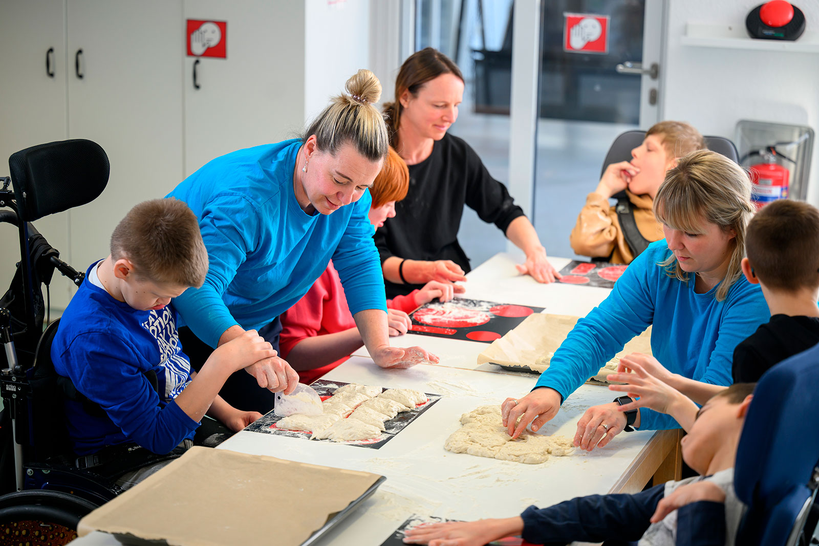 Eine Klasse bereitet gemeinsam Brötchenteig zu: Kinder und Erwachsene kneten und formen Teig an einem großen Tisch.