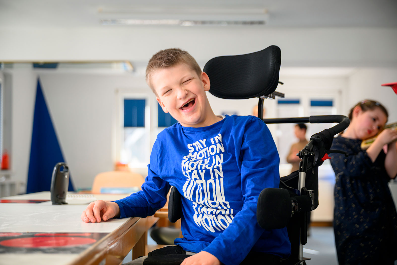 Lucas sitzt im Rollstuhl am Tisch im Klassenzimmer und lacht.