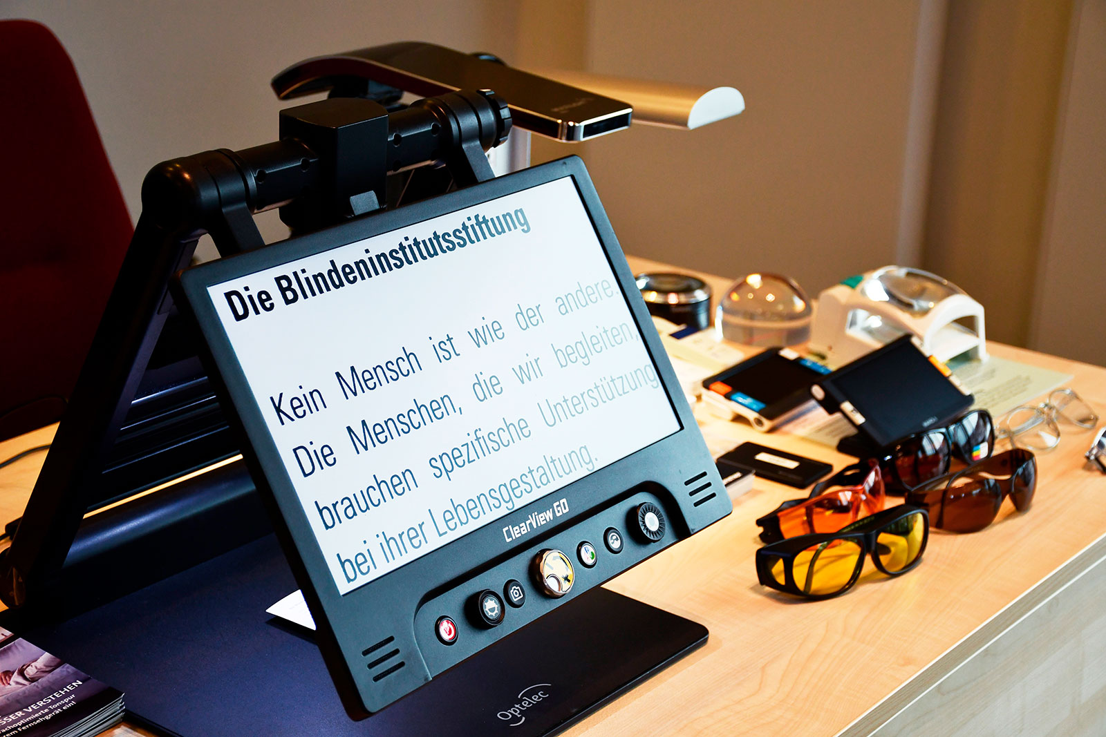 Ein Bildschirmlesegerät zeigt stark vergrößerten Text („Die Blindeninstitutsstiftung“) auf einem Tisch mit Lupen, Lampen, Lesegeräten und mehreren Brillen.