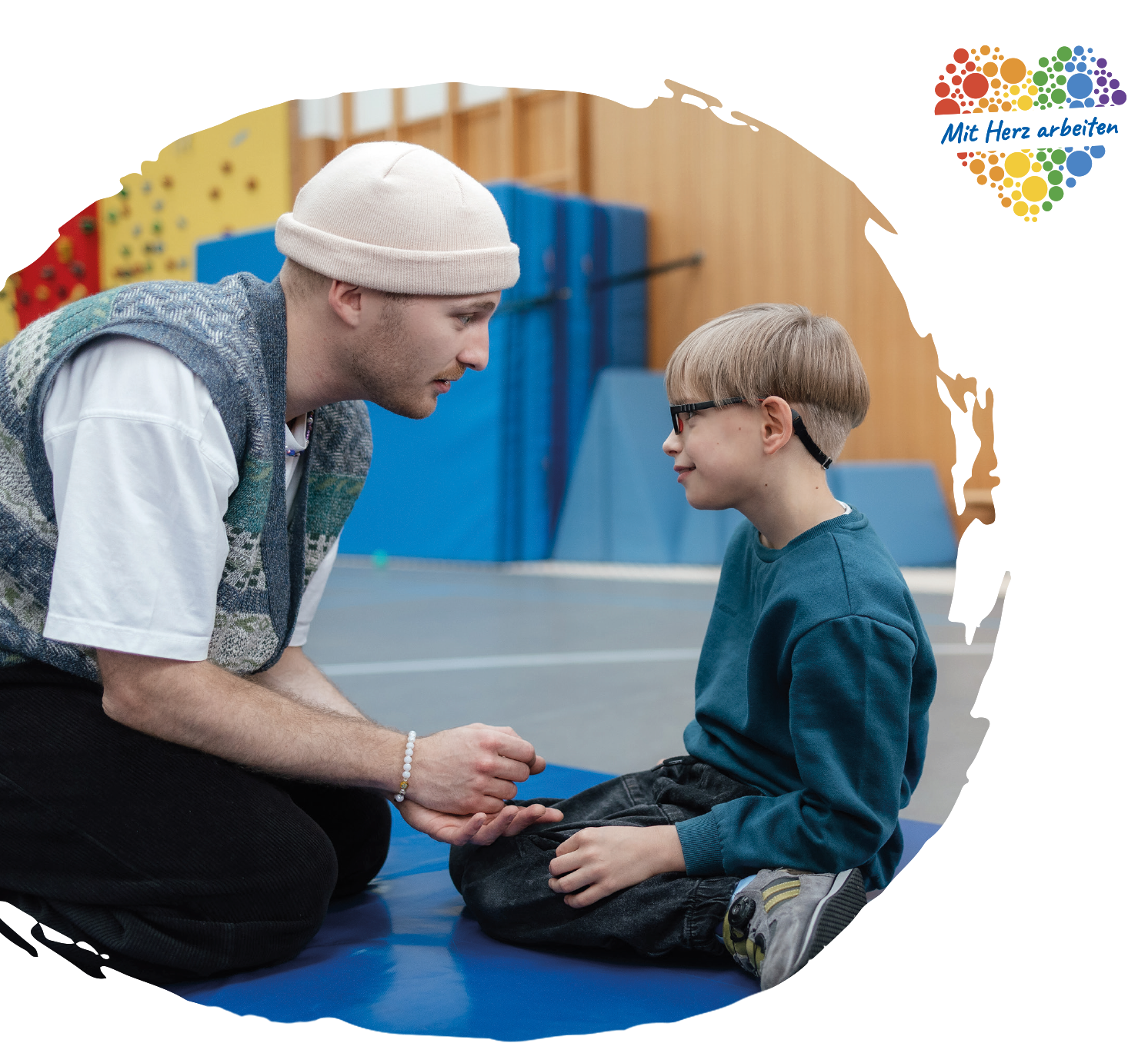 Ein Mitarbeiter des Blindeninstituts spricht in einer Turnhalle mit einem Jungen und sitzt ihm auf Augenhöhe gegenüber; oben rechts steht „Mit Herz arbeiten“.