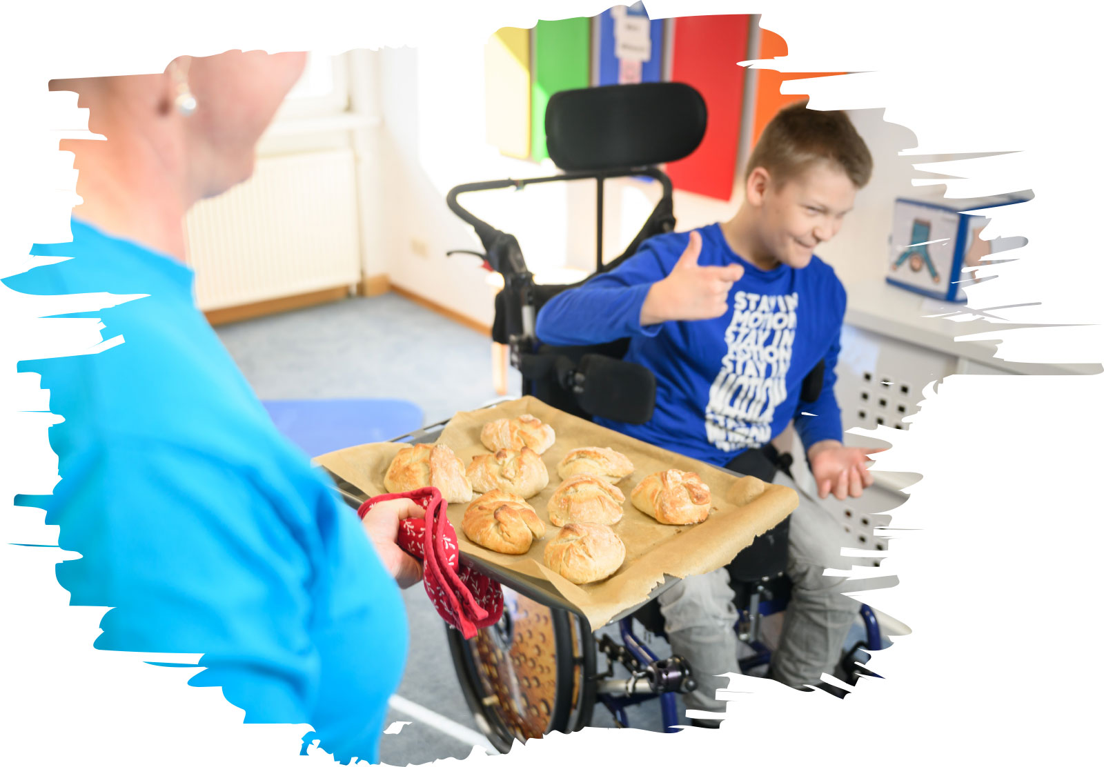 Eine Person hält ein Backblech mit Brötchen; Lucas sitzt im Rollstuhl daneben und zeigt erfreut den Daumen nach oben.