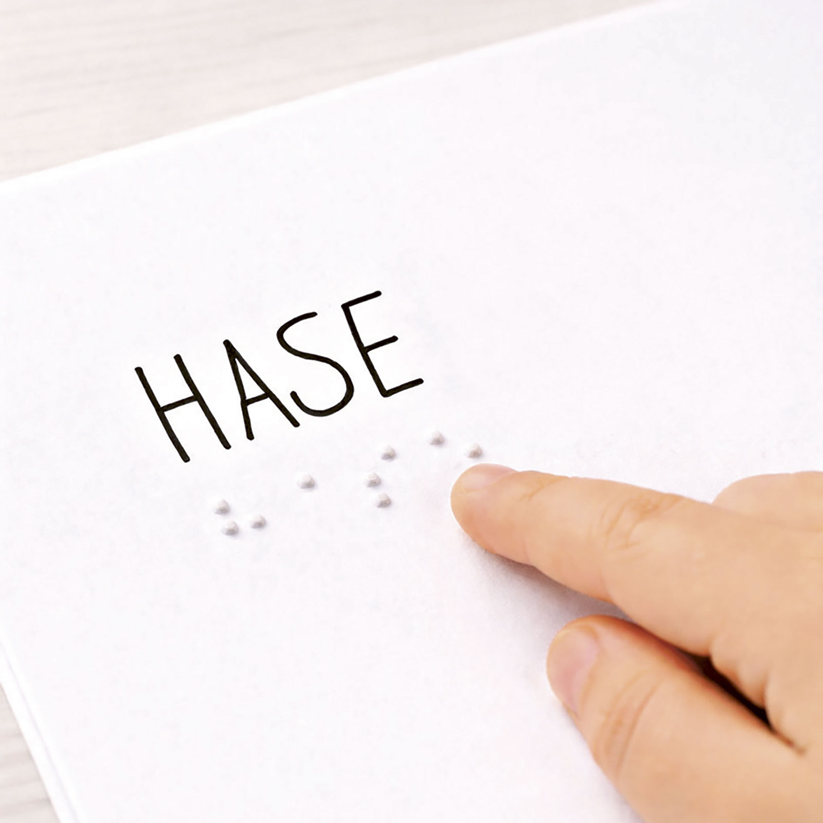 Ein Finger ertastet Braillepunkte auf Papier; darüber steht das Wort „HASE“.