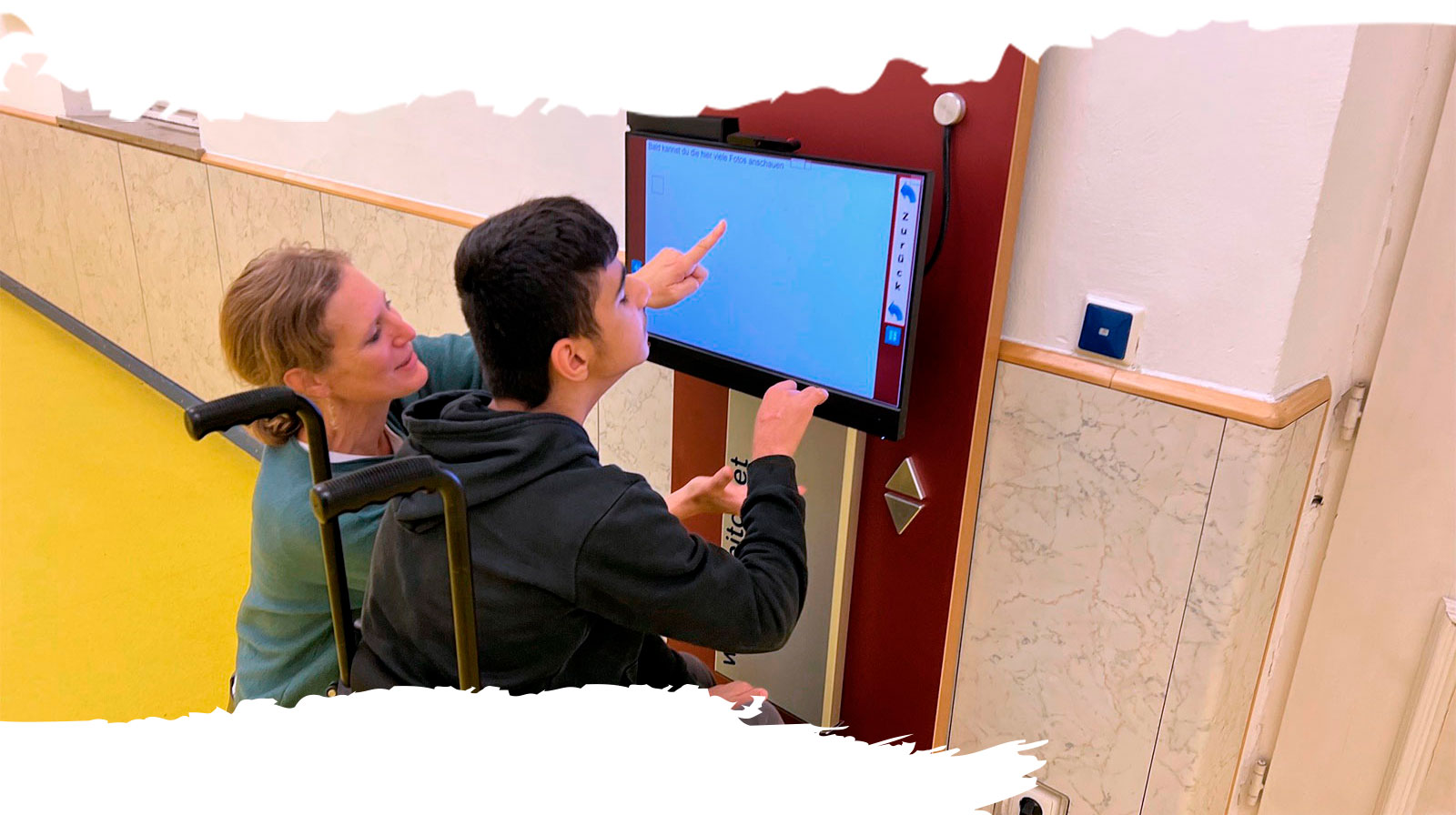 Ein Schüler im Rollstuhl tippt auf den Touchscreen des Informationssystems CABito; eine Mitarbeiterin unterstützt ihn dabei.