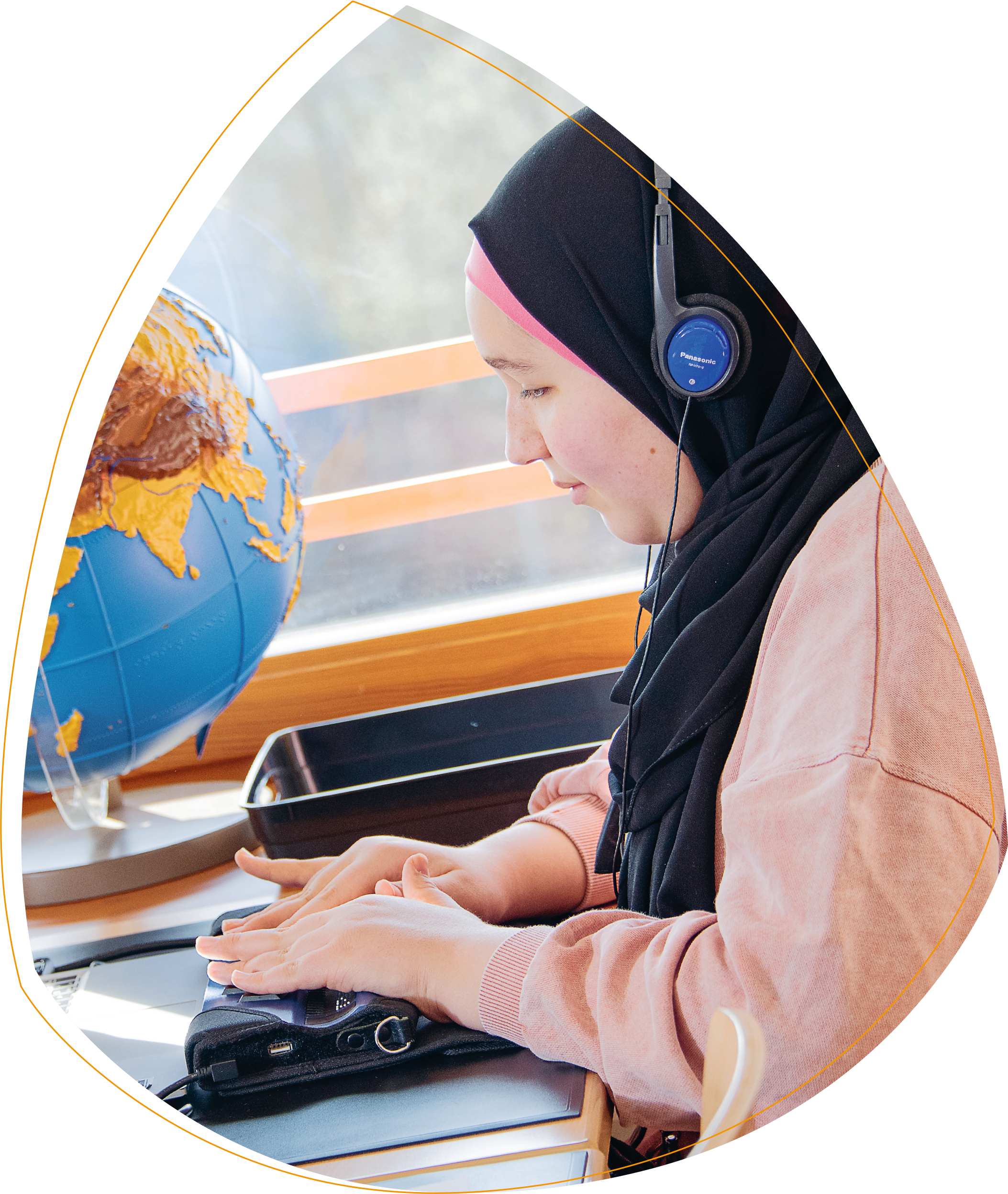 Ein Mädchen mit rosa Pulli und einem schwarzen Kopftuch, das Kopfhörer trägt. Vor ihr liegt eine Braille-Zeile, mit der Sie arbeitet.  Im Hintergrund steht ein Globus, dessen Inhalte physisch hervorgehoben sind. 