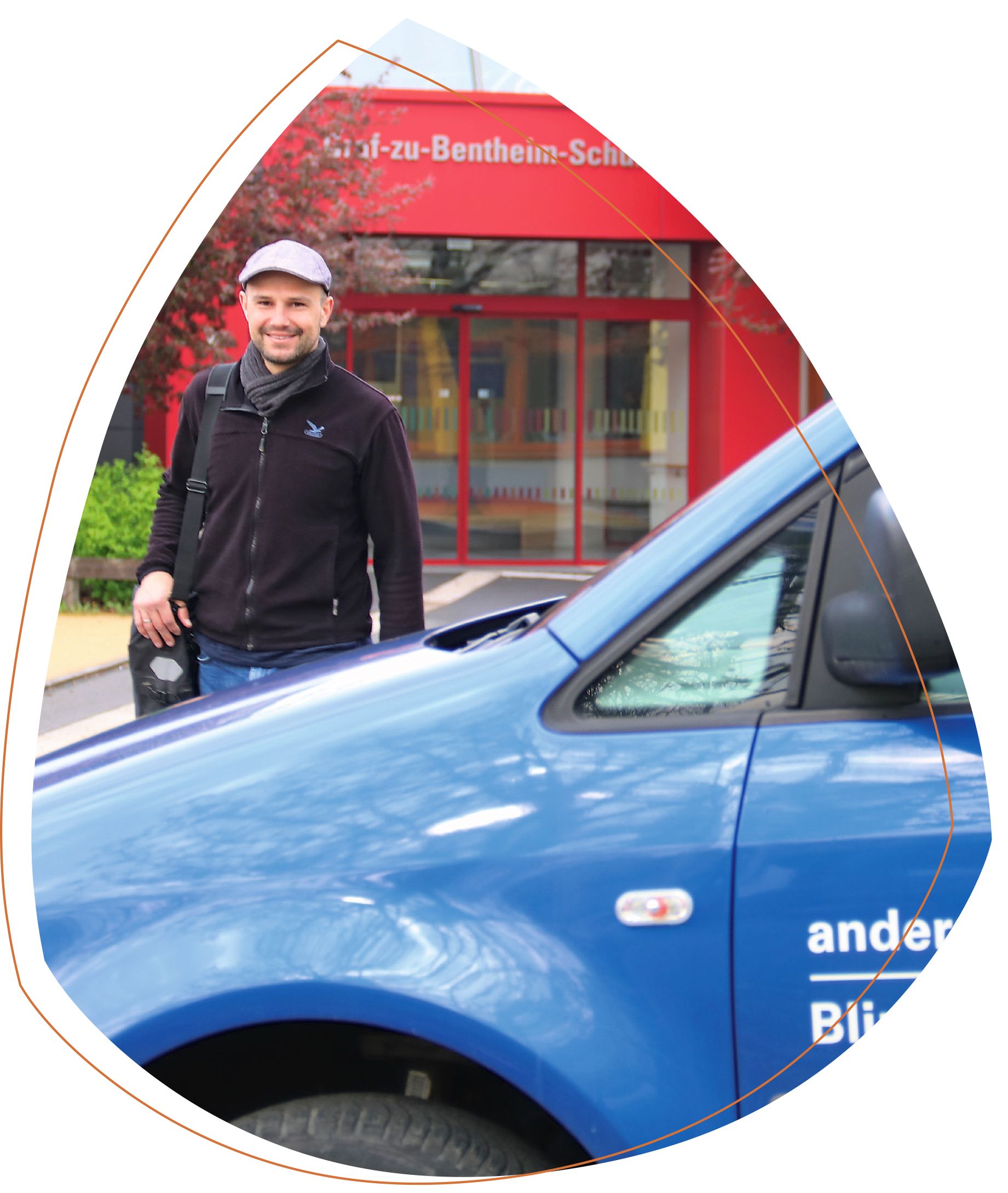Markus Held hinter einem blauen Auto der Blindeninstitutsstiftung. Er trägt eine schwarze Jacke und eine graue Mütze. Auf der linken Schulter trägt er eine Tasche. Im Hintergrund ist die rote Fassade der Graf-zu-Bentheim-Schule zu sehen. 