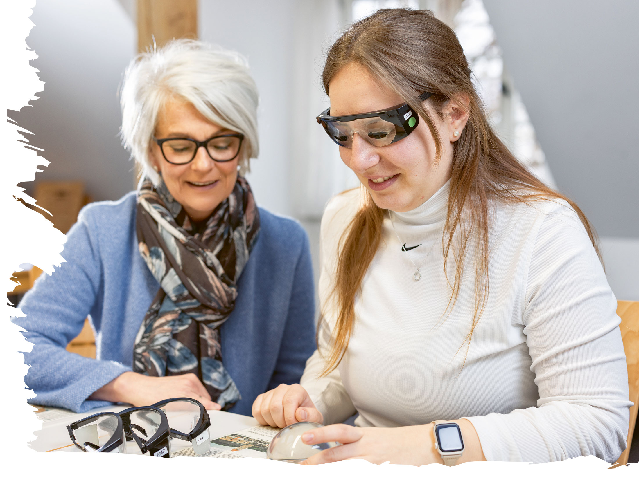 Augenoptikerin Barbara Beitelstein demonstriert einer Pflegekraft, was Menschen mit Sehbeeinträchtigungen tatsächlich sehen. Sie sitzen an einem Tisch, die Pflegekraft trägt eine Brille die eine Sehbeeinträchtigung nachahmen soll.