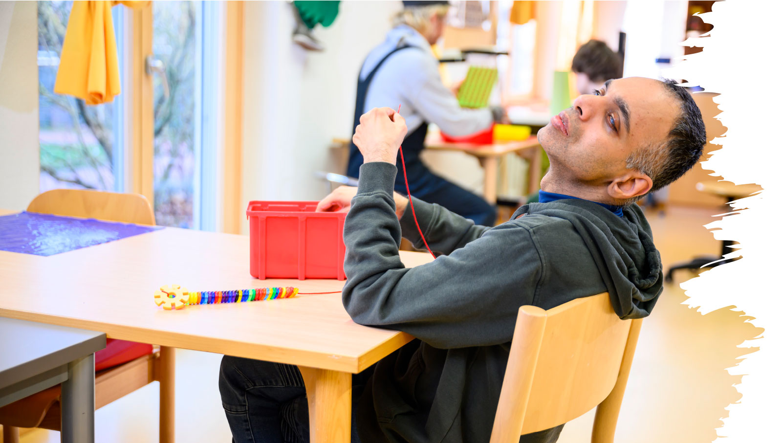 Chinmaya „Chinu“ Kirloskar sitzt an einem Tisch in der Förderstätte und fädelt bunte Perlen auf eine rote Schnur; vor ihm steht ein roter Kunststoffkasten.