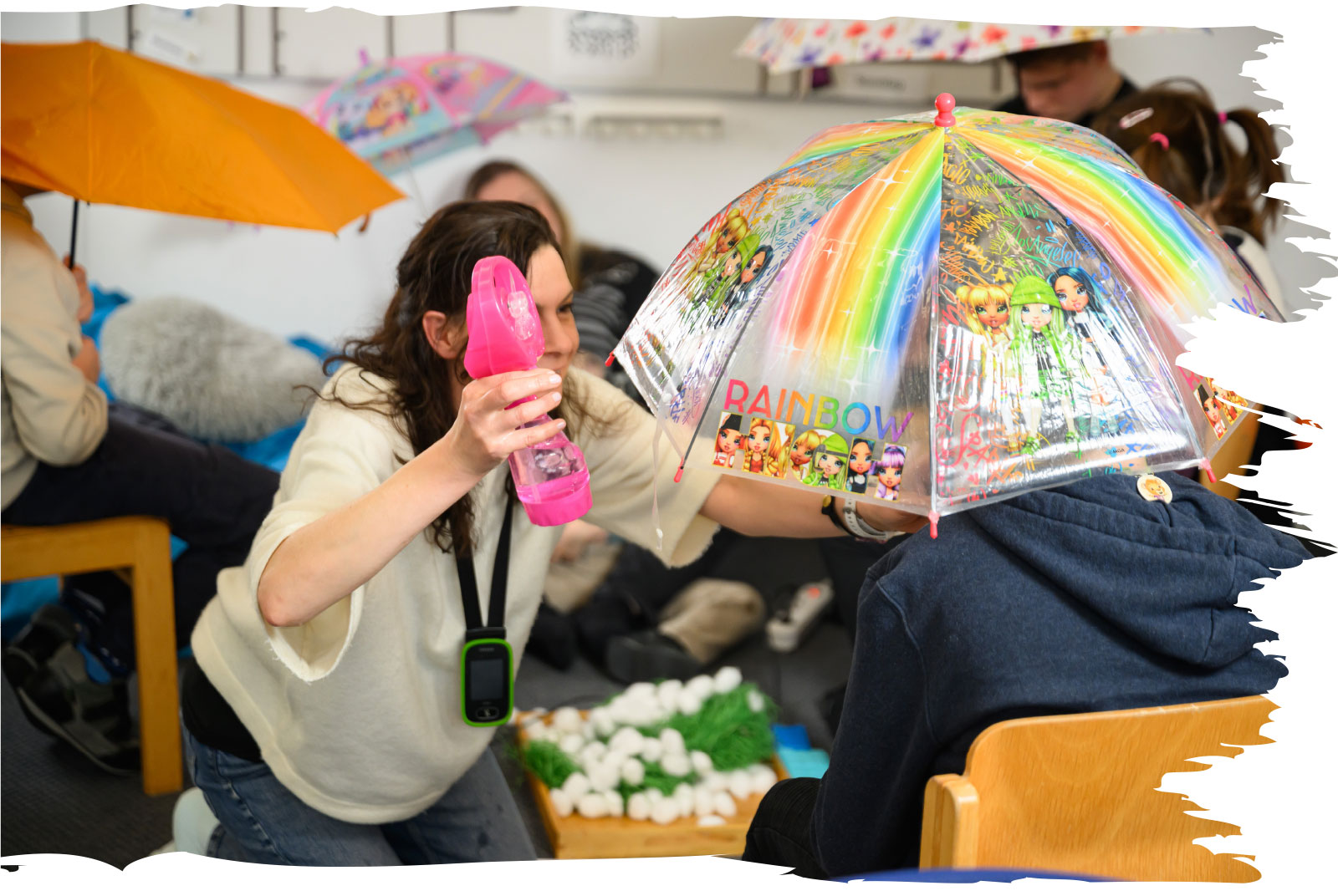 Lehrerin Anja Dantinger sprüht mit einer Sprühflasche Wasser auf einen bunten Regenschirm, den ein Schüler hält.