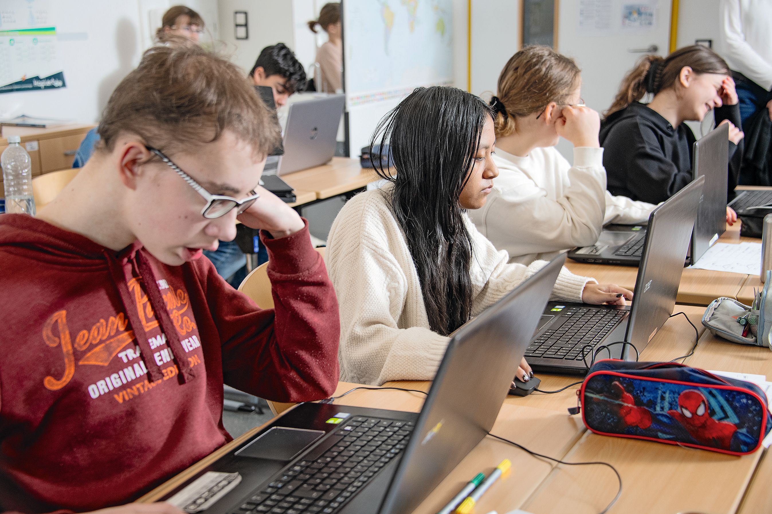 Ein Klassenzimmer in den mehrere Schülerinnen und Schüler sitzen. Alle arbeiten an Laptops. Im Vordergrund sitzt ein junge mit roten Pulli, Brille und hellen Haaren. Neben diesem ein Mädchen mit dunkeln langen Haaren. Sie trägt einen weißen Pulli. 