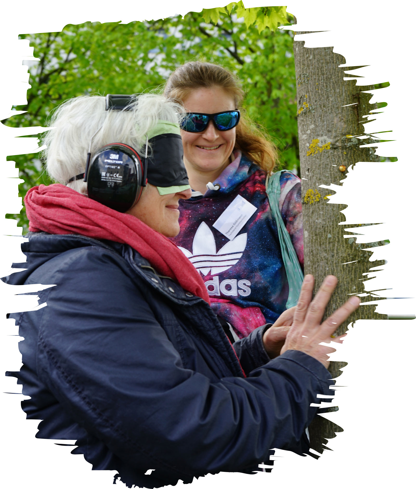 Bei einer Selbsterfahrungsübung trägt eine Person Augenbinde und Gehörschutz und tastet im Freien einen Baumstamm ab; eine Begleitperson steht daneben.