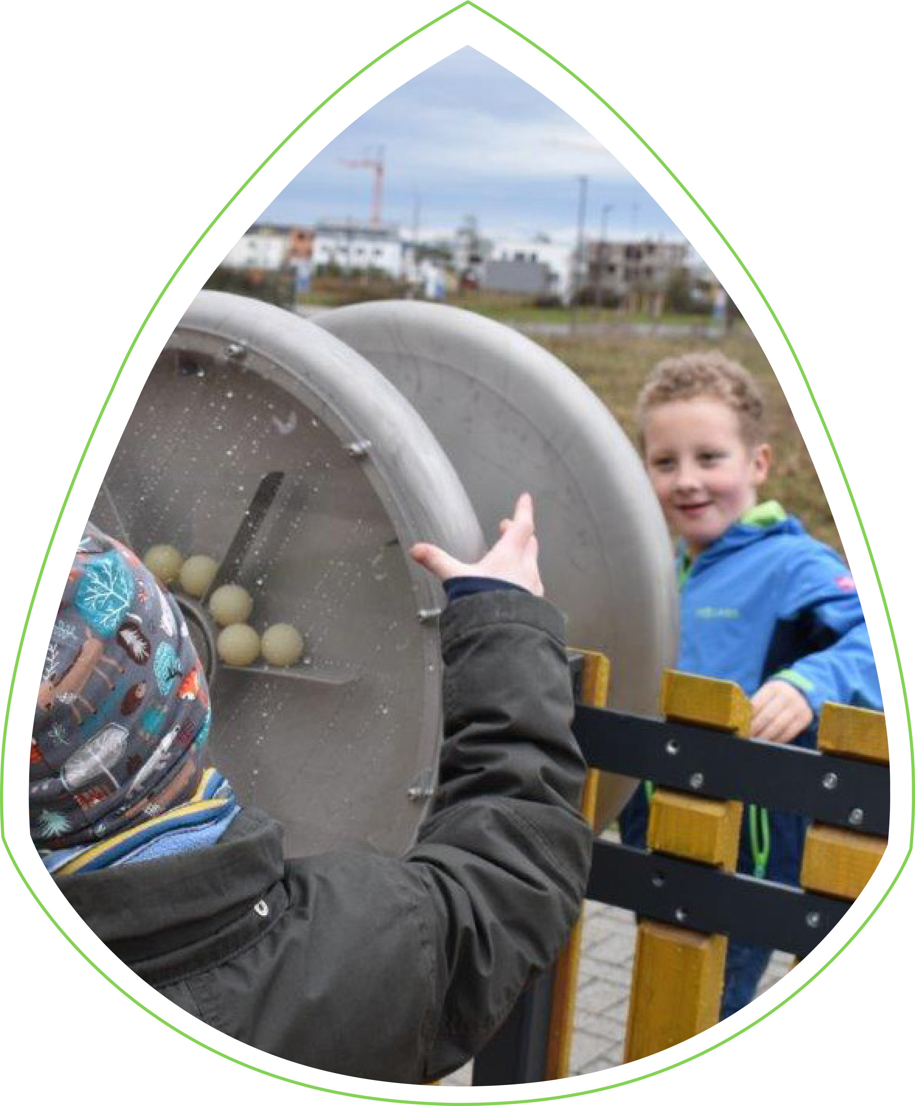 Zwei Kinder spielen am interaktiven Zaun. Hinter dem Zaun steht ein Junge mit einer blauen Jacke. Vor dem Zaun ein Kind, das eine Mütze mit Tier-Muster trägt. Beide drehen an einem Rad, in dem sich weiße Bälle drehen. 