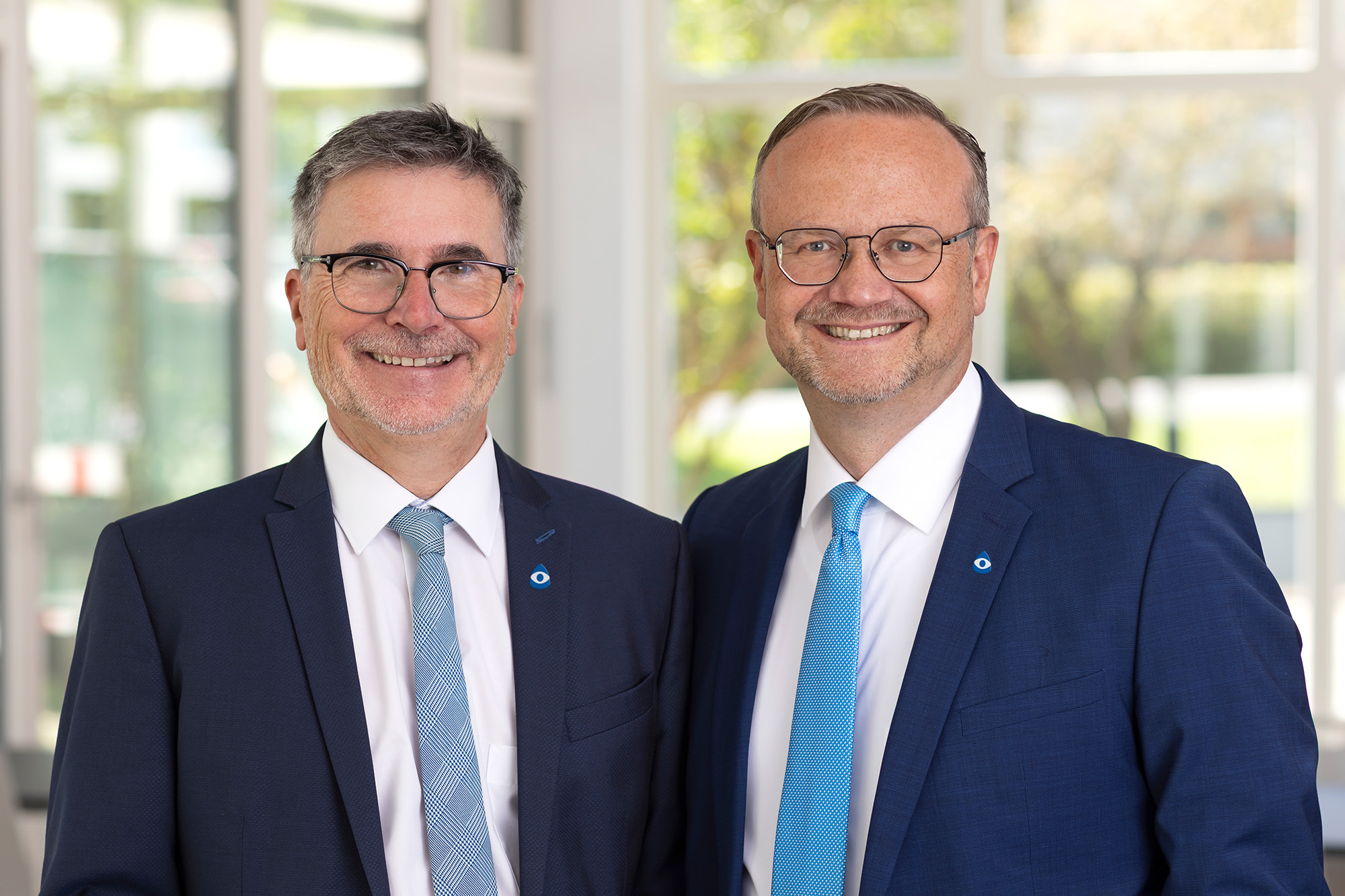 Zwei Männer im dunklen Anzug mit Krawatte lächeln in die Kamera. Sie tragen beide eine Brille.