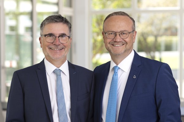 Zwei Männer im dunklen Anzug mit Krawatte lächeln in die Kamera. Sie tragen beide eine Brille.