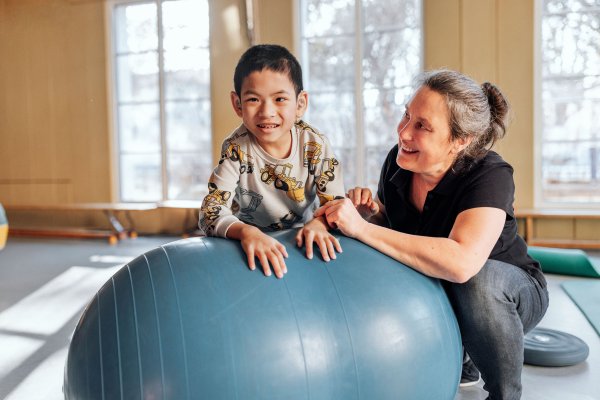 Ein Junge mit Hörgeräten lehnt sich lächelnd über einen großen blauen Gymnastikball. Neben ihm kniet eine Frau in schwarzem Shirt und hält seine Hand. Im Hintergrund ist ein heller Raum mit großen Fenstern und weiteren Turngeräten zu sehen.