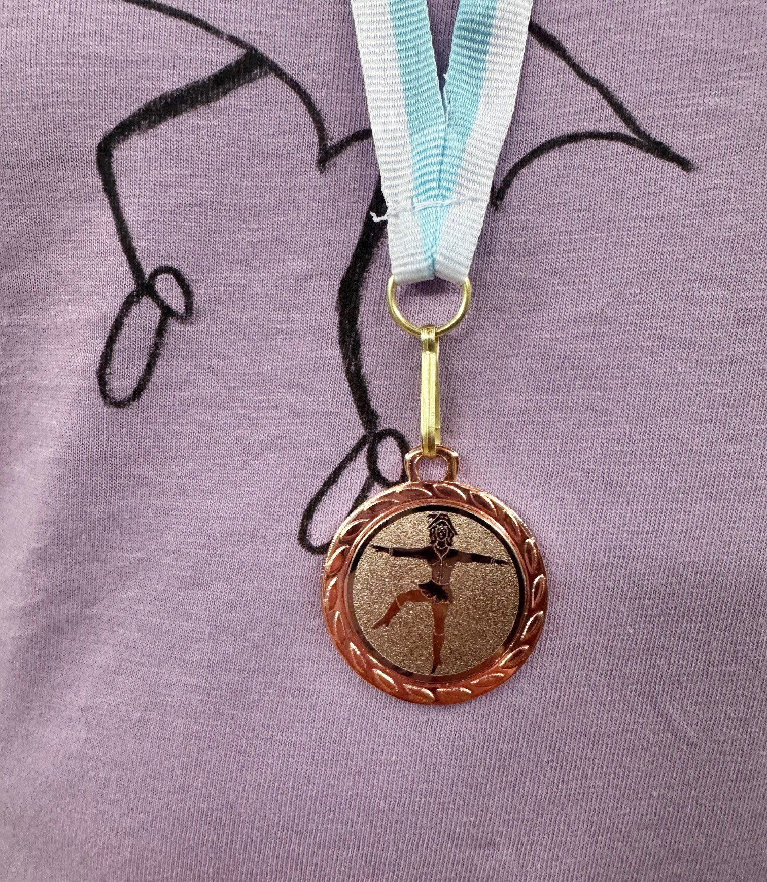 Eine bronzefarbene Medaille mit blau-weiß gestreiftem Band, auf der ein tanzendes Mädchen zu sehen ist.