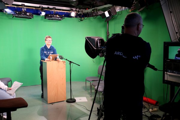 Ein Junge steht im Fernsehstudio vor einem Rednerpult. Vor ihm steht ein Kameramann mit Kamera.