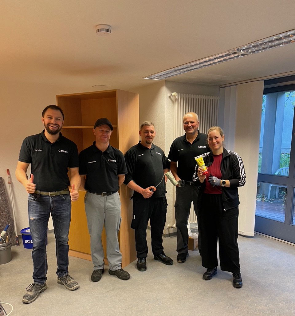 Fünf Personen stehen im einem Halbkreis in einem leeren Raum mit Malerwerkzeugen in der Hand.