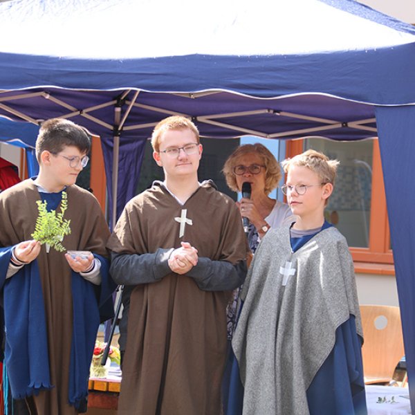 Eine Gruppe von fünf Menschen steht unter einem Pavillon. Links steht der Würzburger Bischof, daneben drei Jungen als Missionare verkleidet und dahinter eine Frau mit Mikrofon.
