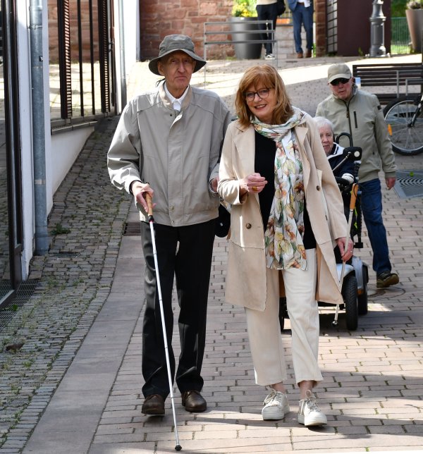 Eine Frau und ein Mann laufen auf einem gepflasterten Weg. Der Mann nutzt einen Langstock, und ist im Arm der Frau eingehakt. Im Hintregrund sind weitere Personen zu sehen, eine von ihnen sitzt in einem elektrischen Rollstuhl.