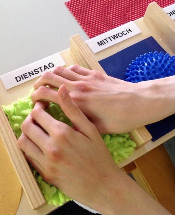 Hände erfühlen einen zotteligen, grünen Waschlappen, der an einem Holzbrett befestigt ist und mit dem Wort Dienstag beschriftet ist.