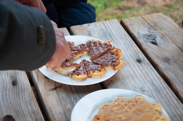 Auf einem Holztisch sind befinden sich Teller mit Waffeln. Eine Waffel ist mit Nutella bestrichen, die andere mit Apfelmus. Jemand schneidet die Waffeln in kleine Stücke.