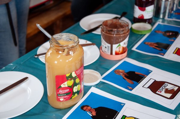 Auf einem Tisch befinden sich Teller, ein Apfelmus-Glas, ein Nutella-Glas und ein Erdbeermarmelade-Glas. Davor liegen ausgedruckte Gebärdenbilder und Metacom-Symbole.