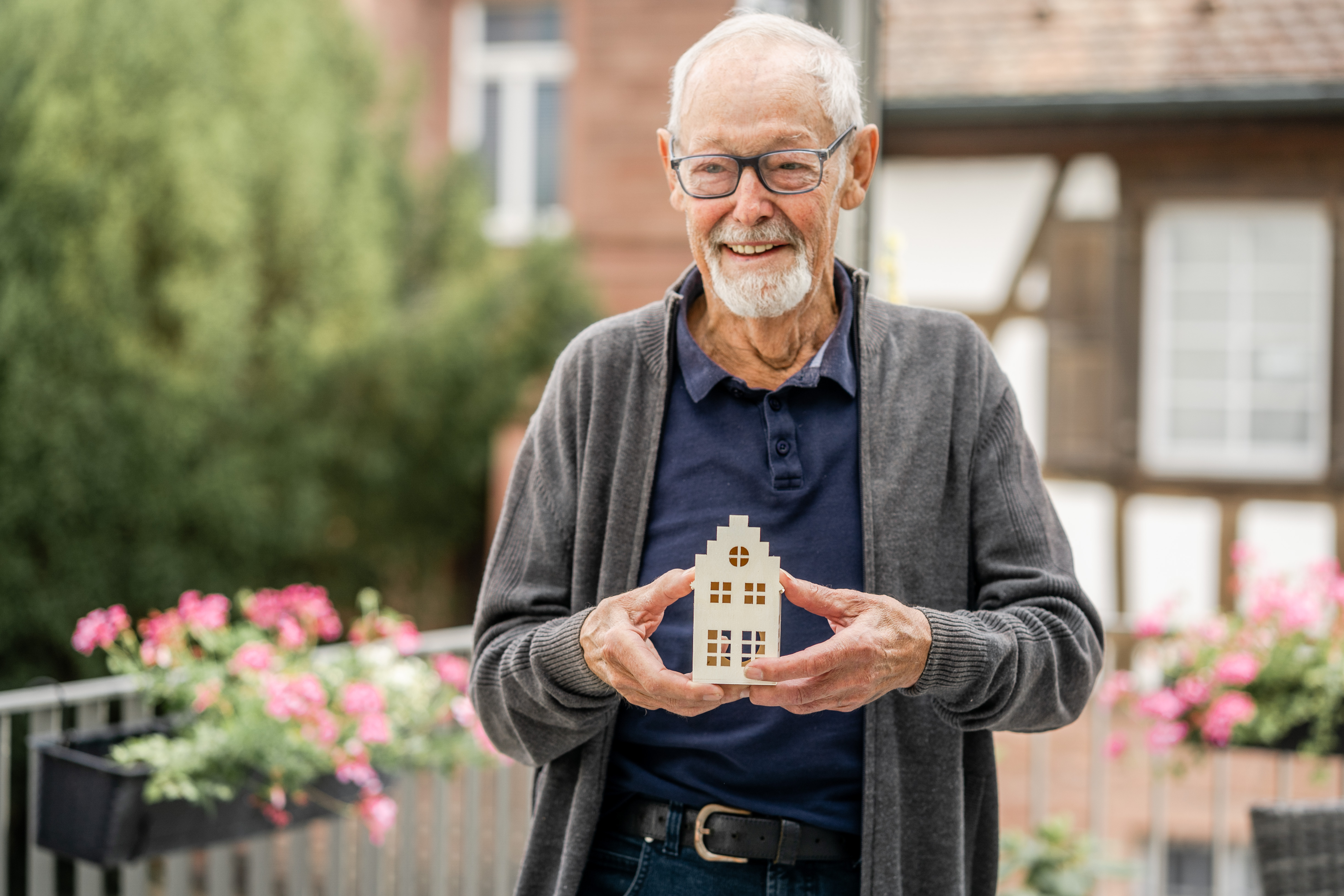 Ein lächelnder Senior hält ein Holzhäuschen in den Händen. Im Hintergrund befindet sich ein Fachwerkhaus. Der Senior steht auf einem Balkon, der mit Blumen geschmückt ist. 