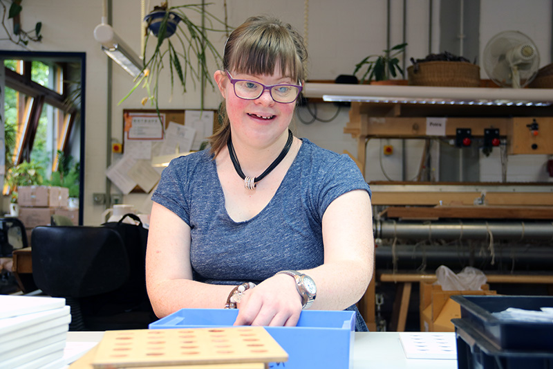 Junge Frau mit Brille sitzt an einem Arbeitstisch, greift in eine blaue Kiste und sortiert kleine Teile; im Hintergrund Werkstattregale und Pflanzen am Fenster.