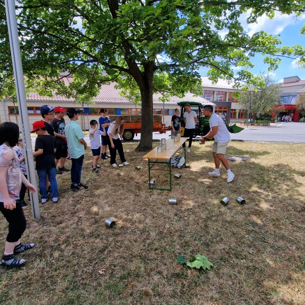 Eine Gruppe Kinder steht links im Halbkreis vor einem Tisch auf dem leere Konservendosen aufgebaut sind. Ein Kind versucht mit einem Ball die Dosen vom Tisch zu werfen. Rechts daneben steht ein erwachsener Mann und beobachtet das Geschehen.