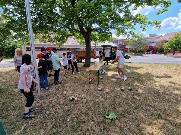 Eine Gruppe Kinder steht links im Halbkreis vor einem Tisch auf dem leere Konservendosen aufgebaut sind. Ein Kind versucht mit einem Ball die Dosen vom Tisch zu werfen. Rechts daneben steht ein erwachsener Mann und beobachtet das Geschehen.