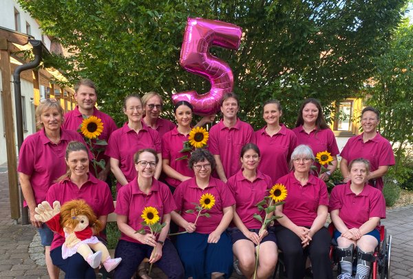 Alle Mitarbeitenden des MZEB sitzen in zwei Reihen hintereinander in der pinken MZEB-Kleidung mit einer Sonnenblume in der Hand. Hinten in der Mitte schwebt ein pinker Luftballon in Form einer Fünf.