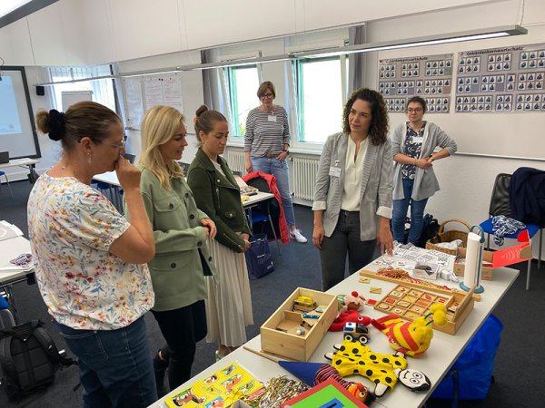 Im Bildvordergrund ist ein Tisch mit leuchtend bunten Frühförderspielzeugen abgebildet, dahinter stehen vier Frauen, die das Spielzeug betrachten und miteinander sprechen. Im Hintergrund beobachten zwei weitere Frauen das Geschehen.