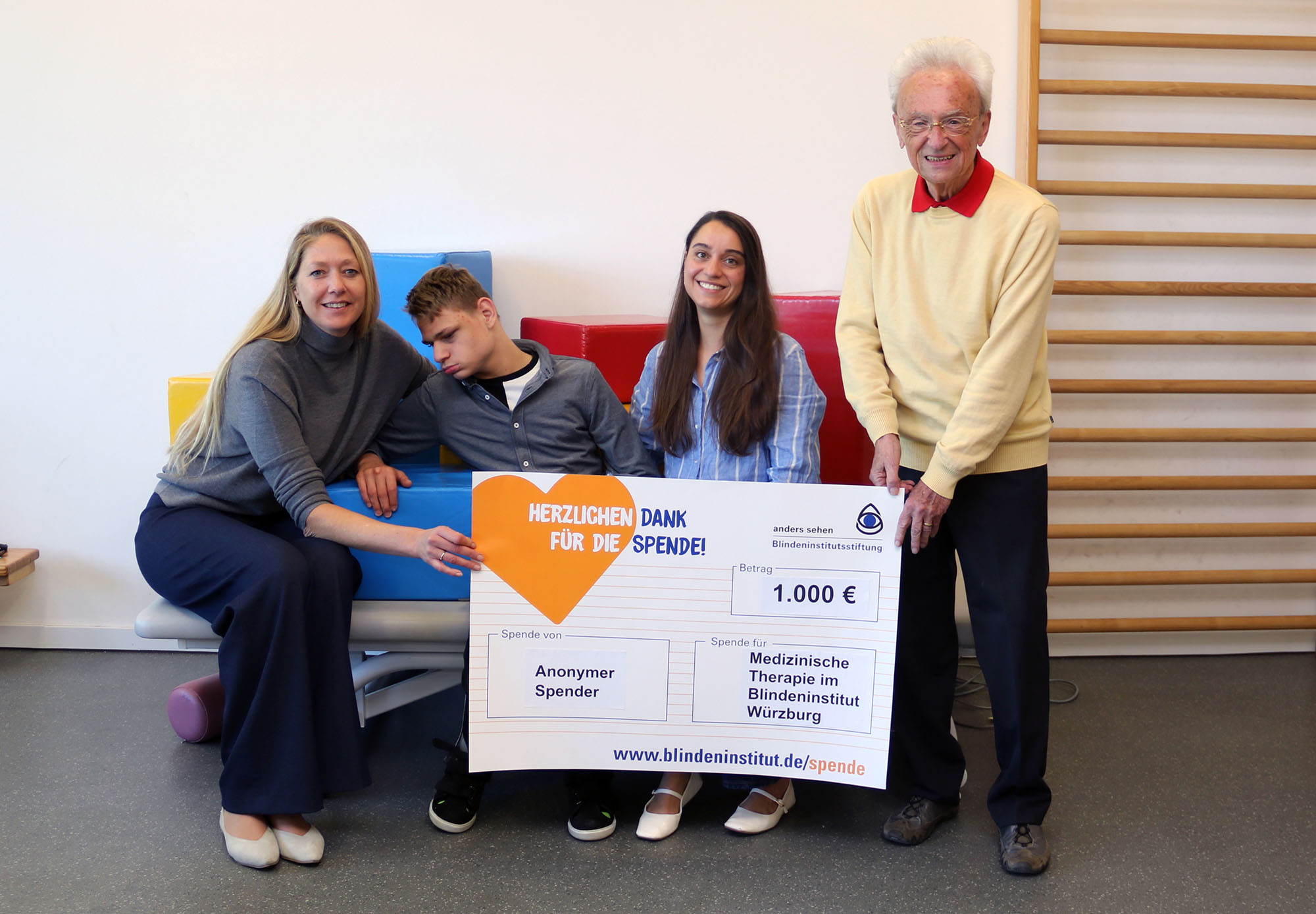 Vier Personen sitzen auf einer Therapieliege und halten einen Spendenscheck in der Hand
