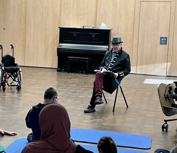 Ein Mann sitzt auf einem Stuhl in einem großen Raum, vor ihm sitzen Kinder auf dem Boden und hören ihm zu.