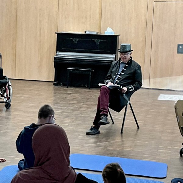 Ein Mann sitzt auf einem Stuhl in einem großen Raum, vor ihm sitzen Kinder auf dem Boden und hören ihm zu.